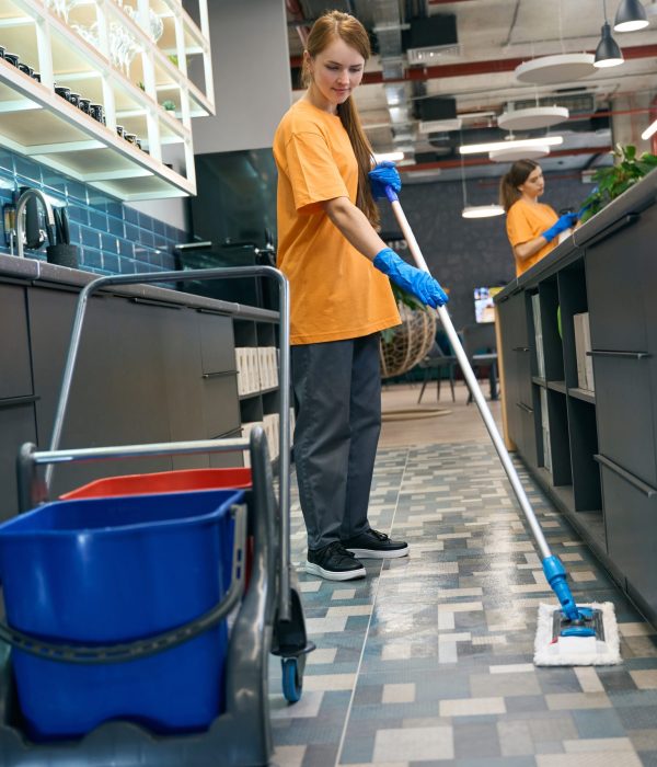 Young woman cleaning the floor with a mop, her colleague is in the background